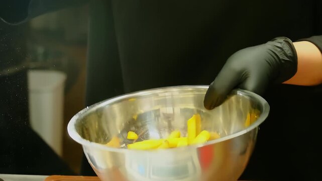 Cook tosses french fries in metal bowl to spread salt. Gloved hand flips bowl upward quickly while fries tumble settling for even seasoning