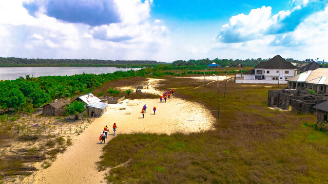 Aerial view of people walking along a sandy path near simple buildings, with a river and lush green trees in the background, Olu Ama, Rivers State, Nigeria.