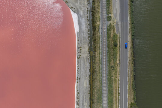 Aerial view of startling contrast between pink salt ponds and green waters running parallel to a road with a lone blue car, Aigues-Mortes, Occitanie, France.
