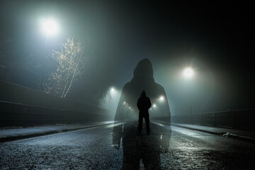 Double exposure of a mysterious hooded figure standing on a road on a creepy foggy winters night.