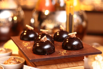Luxury chocolate mirror glaze mousse domes with gold leaf and chocolate garnish. Sophisticated artisanal patisserie served on a wooden board at a high-end restaurant buffet or gala event.