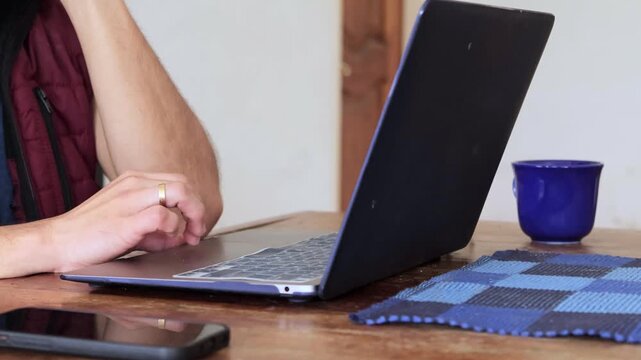 Detail shot of man's hands with a gold wedding ring working on a laptop trackpad and keyboard, sitting at a wooden table with a blue coffee mug and smartphone nearby.