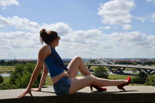 Outdooraufnahme einer sportlich gekleideten Frau von hinten, die von einer erh&ouml;hten Position auf eine Stadtlandschaft mit Fluss und Br&uuml;cke blickt. Blauer Himmel und sommerliches Licht vermitteln Freih