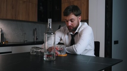 Adult man in a white shirt sitting alone in a dark room and drinking strong alcohol. Empty and half-empty bottles and a glass on the table in front of him. Concept of alcohol addiction