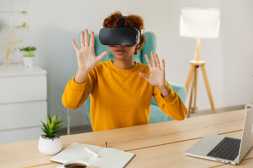 African American young woman wearing using virtual reality metaverse VR glasses headset at home. Girl touching air during VR experience on virtual reality helmet. Simulation hi-tech videogame concept