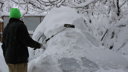 A man in a black jacket and a bright hat with a long-handled scraper clears a car of a huge snowdrift after a snow storm in a private yard. Clearing vehicles after a huge snowfall.