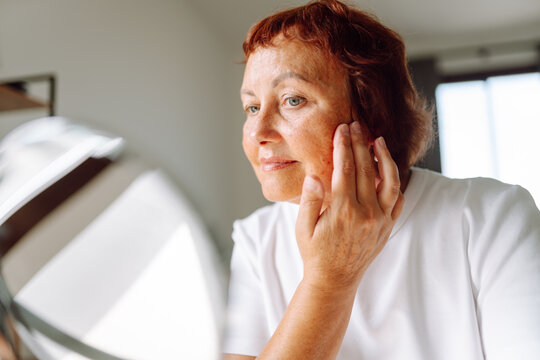 Portrait of adult woman looking into small mirror and touching face while sitting in cozy room. Woman examines wrinkles, enjoys natural beauty indoors on sunny day. Concepts of aging, natural beauty.