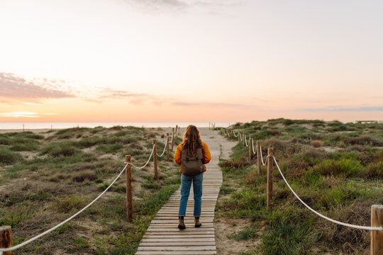 A young woman with a backpack walks along a wooden path to the beach at dawn. The woman, wearing a bright jacket, enjoys the seascape and the hike. Adventure, hiking concept.