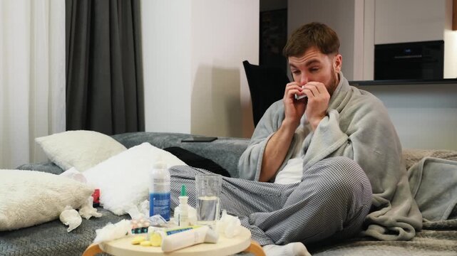Sick man suffering from a cold wrapped in blanket sitting on sofa in living room at home. A tired bearded male coughs, sneezes, blows his nose with a handkerchief. He has fever or flu.