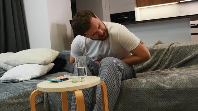 Unhappy man, sits on comfortable sofa in cozy living room, holds his hands on his stomach, grimaces from pain in his stomach, suffers from poisoning, spasm, stomach problems