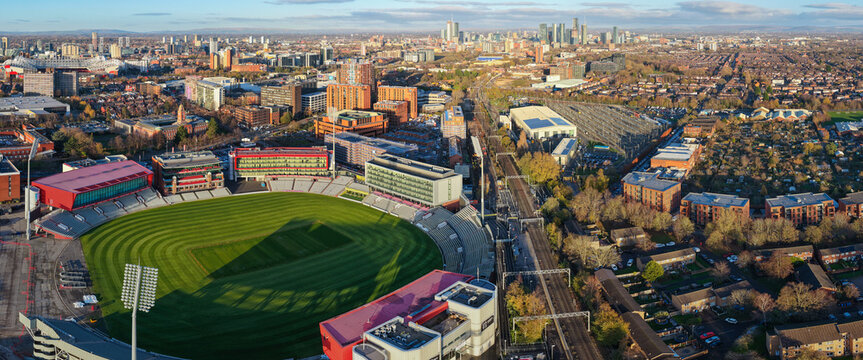 A wide-angle panoramic aerial view of the Emirates Old Trafford Cricket Ground, looking toward the Manchester United football stadium and the city center skyline at sunset.
