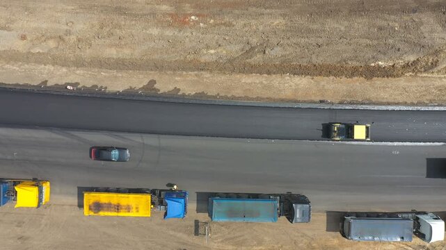 Aerial shot of roller making new asphalt road and trucks standing at roadside. Special machinery repairing roadway in countryside. Drone tracking compactor constructing highway. Top view