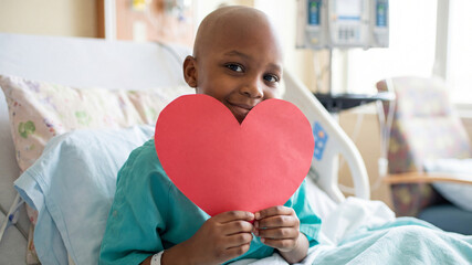Bald African American Child with red paper heart in hospital bed. Concept of health, hope, recovery for Valentine's Day or childhood cancer with copy space.