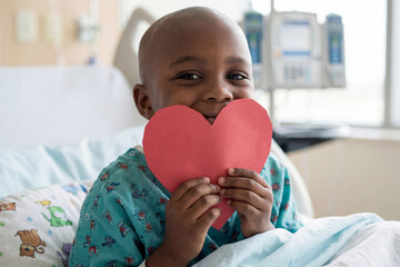Bald African American Child with red paper heart in hospital bed. Concept of health, hope, recovery for Valentine's Day or childhood cancer with copy space.