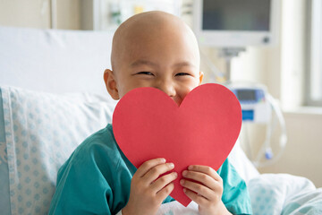 Bald Child asian patient with bald head holding red heart card in hospital bed. Oncology pediatric patient with cancer for compassionate care, recovery for Valentine's Day or childhood cancer.