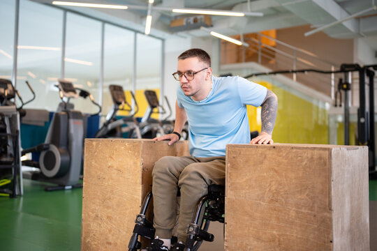 Dedicated disabled man with spinal injury recovering at gym center doing adaptive workout with simple equipment. Male athlete in wheelchair working on arms and upper body strength using wooden blocks.