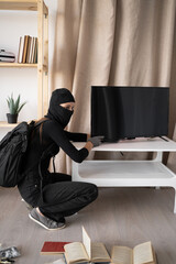 Woman burglar or thief in black balaclava stealing tv set from house. Robber crime