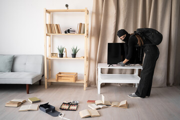 Woman burglar in black balaclava stealing tv set from house. Robber terrorism