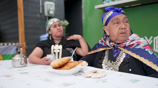 Two indigenous women in traditional mapuche clothing sit at a table, drinking mate and talking