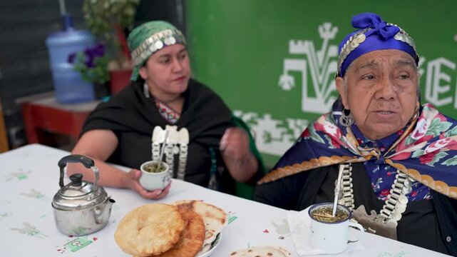 Two native mapuche women in traditional attire drinking mate and eating sopaipillas at a table