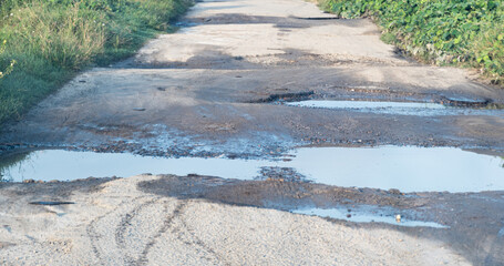 Large water-filled potholes and puddles disrupt the surface of a weathered rural road during daylight hours.