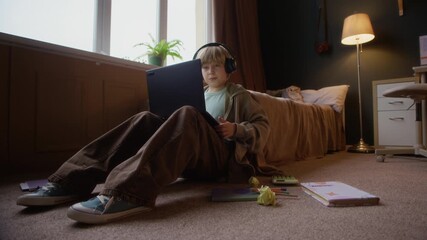 Ground level shot of cheerful Caucasian student boy sitting on floor in bedroom in front of laptop surrounded by educational materials and waving at screen having online lesson with tutor