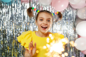 Happy smiley girl in glasses and yellow holiday blouse with ponytails hairstyle on festive background with balloons looks at sparklers