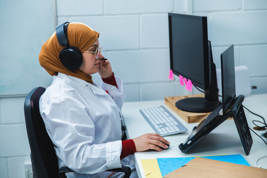 Muslim female scientist in hijab and lab coat working on computer and wearing headset at office desk
