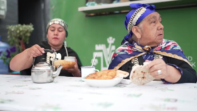 Two indigenous women in traditional clothing enjoying sopaipillas and mate tea at an outdoor table