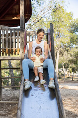 Obraz premium Joyful mother and toddler sliding down a slide at a forest park