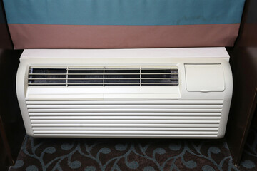 Hotel room air conditioner unit under window. White wall mounted HVAC system front view. Cooling and heating appliance indoors. Climate control equipment in modern interior. Energy use concept.