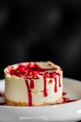 A mini white chocolate raspberry cheesecake on a white plate with a dark background. Features a graham cracker crust, creamy filling, red sauce drizzle, and freeze-dried berry crumbles.