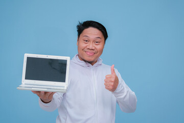 closeup of a young adult man raising his thumb in front of his laptop while laughing broadly like an advertisement wearing white t-shirt on turquoise blue  