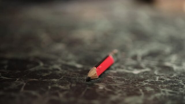 Macro shot of a tiny, used red pencil on a dark textured surface. The focus shifts, highlighting the worn wood and graphite tip of this small writing tool.

