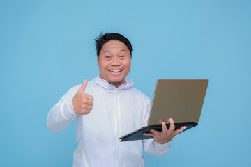 closeup of a young adult man raising his thumb in front of his laptop while laughing broadly like an advertisement wearing white t-shirt on turquoise blue  