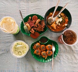 Indonesian Street Food Feast with Takoyaki and Iced Drinks. The meal includes a plate of takoyaki topped with seaweed, a variety of deep-fried snacks (gorengan), a bowl of spicy noodle soup or seblak,