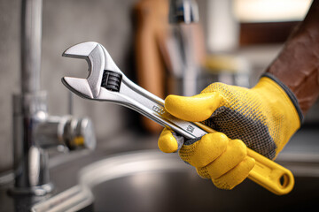A hand in a yellow glove holds a silver adjustable wrench, ready for plumbing work near a sink in a well-lit room.