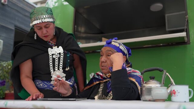 Two indigenous mapuche women in traditional clothing signing a document on a digital tablet