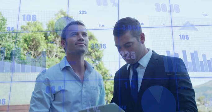 Reviewing charts, two men in light blue shirt and dark suit studying tablet by glass wall - Powered by Adobe