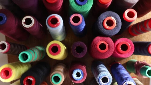 Top down view of many colorful spools of sewing thread on a wooden surface. Camera pans over the vibrant collection of tailoring materials and textile supplies.

