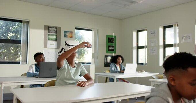 African American students seeing youth female in white VR headset raising arm for teacher attention