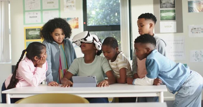 Diverse teens gathering in class watching African teen putting on VR headset and tapping tablet