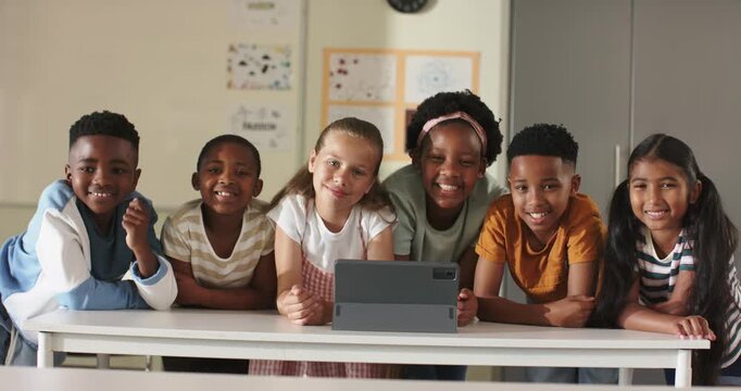 Six diverse schoolchildren leaning on table in classroom, peering at tablet on stand and smiling