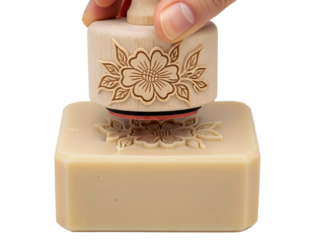 Wooden stamp pressing floral pattern into beige handmade soap bar on a transparent background