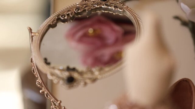 Rack focus shot of an ornate vintage mirror. The focus shifts from the decorative gold frame to a pink flower reflection, evoking a romantic and elegant atmosphere.

