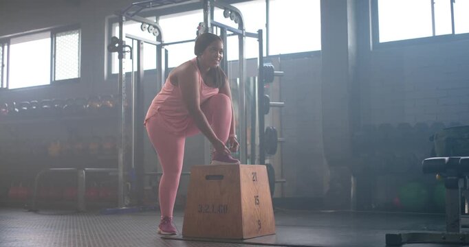 African American woman in pink approaching plyo box at gym, placing foot tying laces for step-up