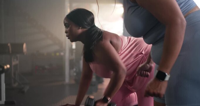 Female partners performing one-arm dumbbell rows on bench at gym, partner approaching guiding form