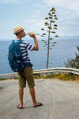 Teenage boy drinking water from a plastic bottle during a summer hike with sea view and a flowering agave in the background. Outdoor adventure, hydration, and scenic coastal exploration.