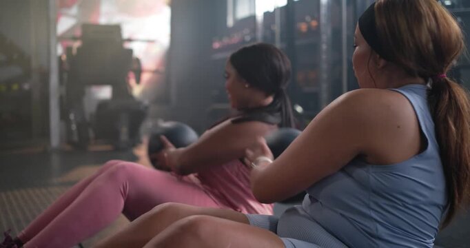 Diverse female friends leaning back, holding black medicine balls, rotating torsos for core at gym