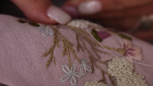 Close-up view of female hands carefully embroidering delicate white flowers onto pink fabric. The detailed needlework features floral patterns and lace edges, showcasing traditional craftsmanship and 